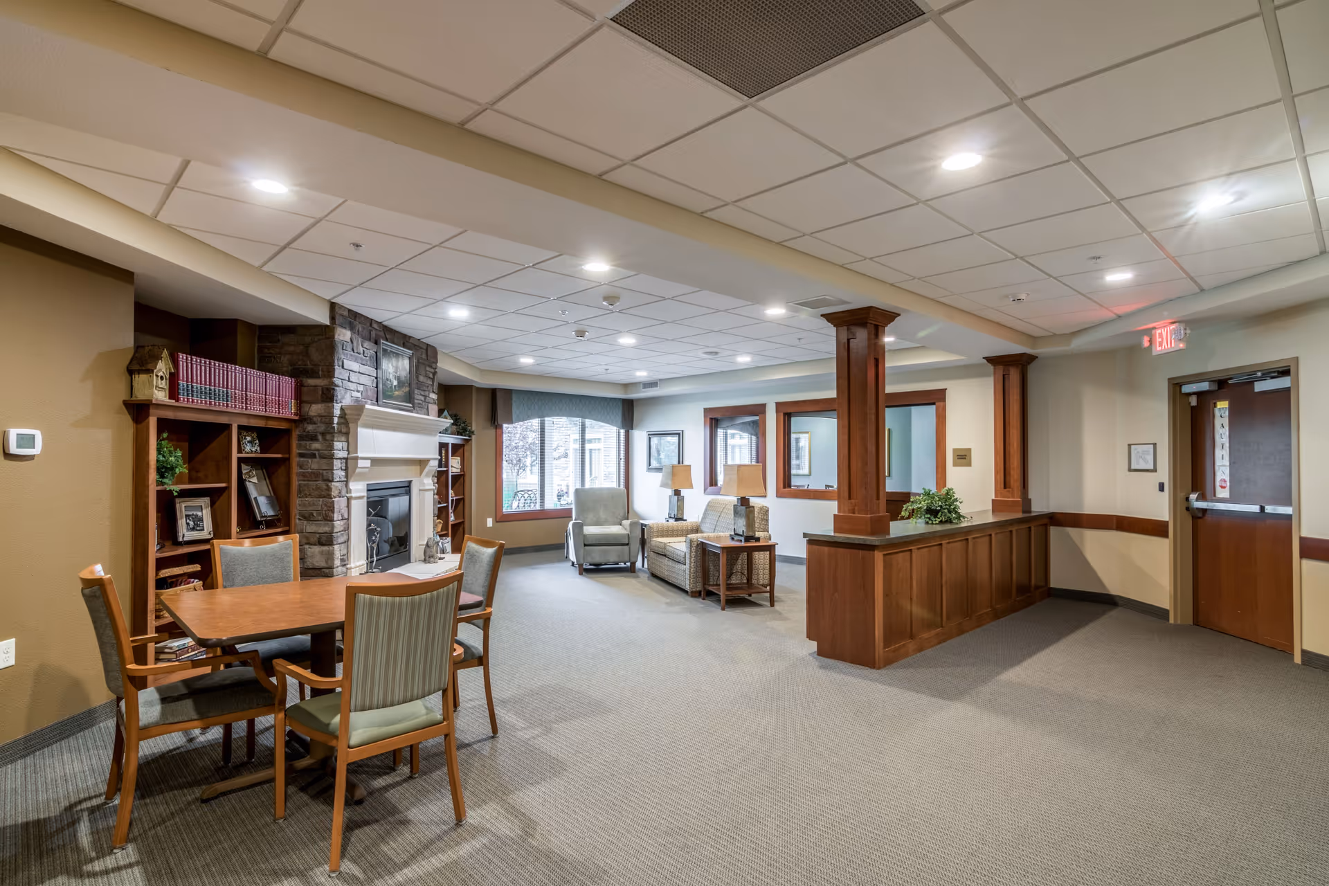 A spacious senior living common area with a stone fireplace, wooden bookshelves filled with books and framed photos, a wooden table with four chairs, two armchairs, a sofa, side tables with lamps, and large windows letting in natural light. The room features beige walls, carpeted floors, and a drop ceiling with recessed lighting.
