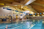 Indoor lap swimming pool with lane lines, overhead wooden beams, and swimmers in the water.