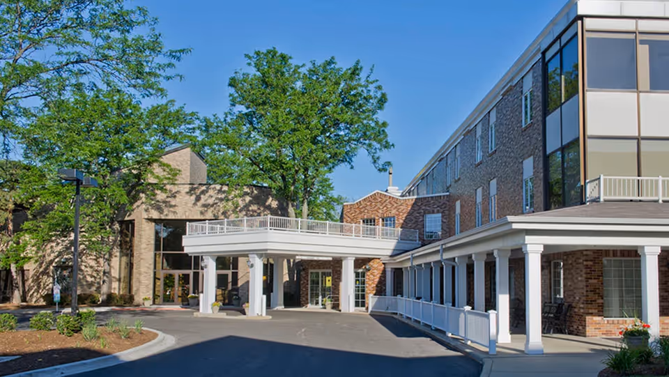 Exterior view of a multi-story senior living facility building with brick walls, large windows, a covered entrance supported by white columns, and surrounding trees under a clear blue sky.