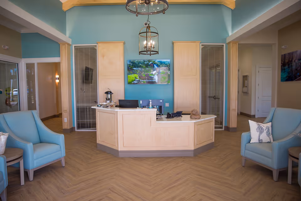 Reception area of A Banyan Residence Assisted Living Resort with a light wood reception desk, two light blue armchairs with decorative pillows, a wall-mounted TV, and pendant lighting. The walls are painted in a soft blue and beige color scheme, and the floor has a wood pattern.