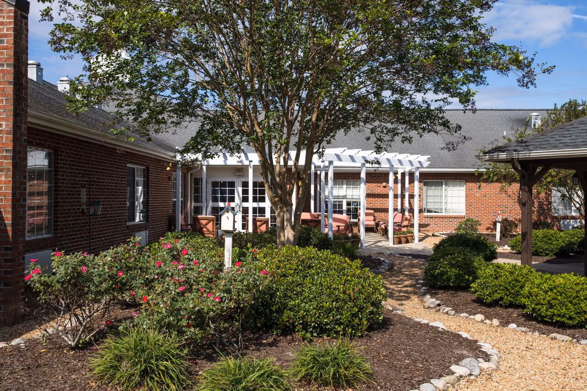 Landscaped courtyard with flowering bushes, a central tree, gravel paths and patio seating under a white pergola in front of a brick building.