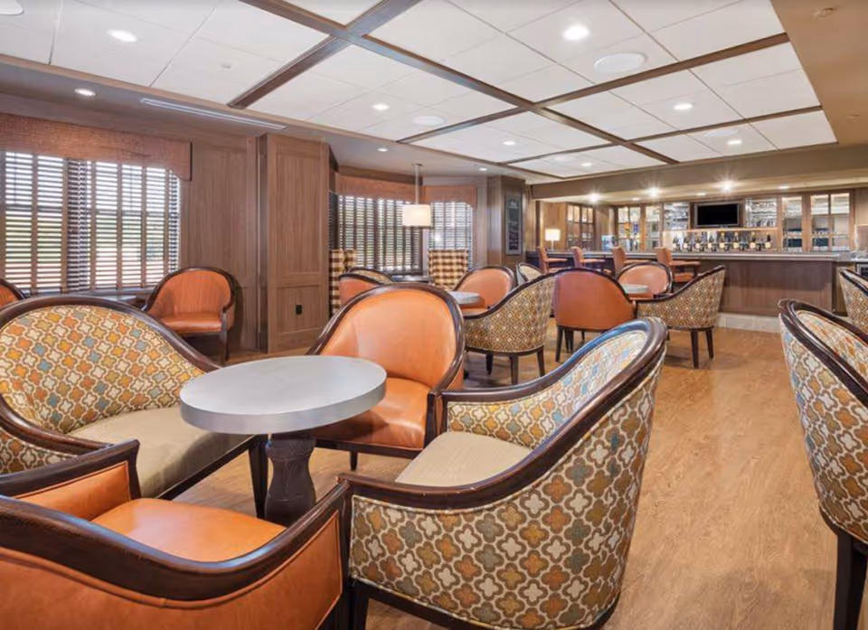 A spacious senior living common area with multiple seating arrangements including patterned armchairs and orange leather chairs around small round tables. The room features wood paneling, large windows with blinds, and a bar area with high chairs and a television mounted above it.