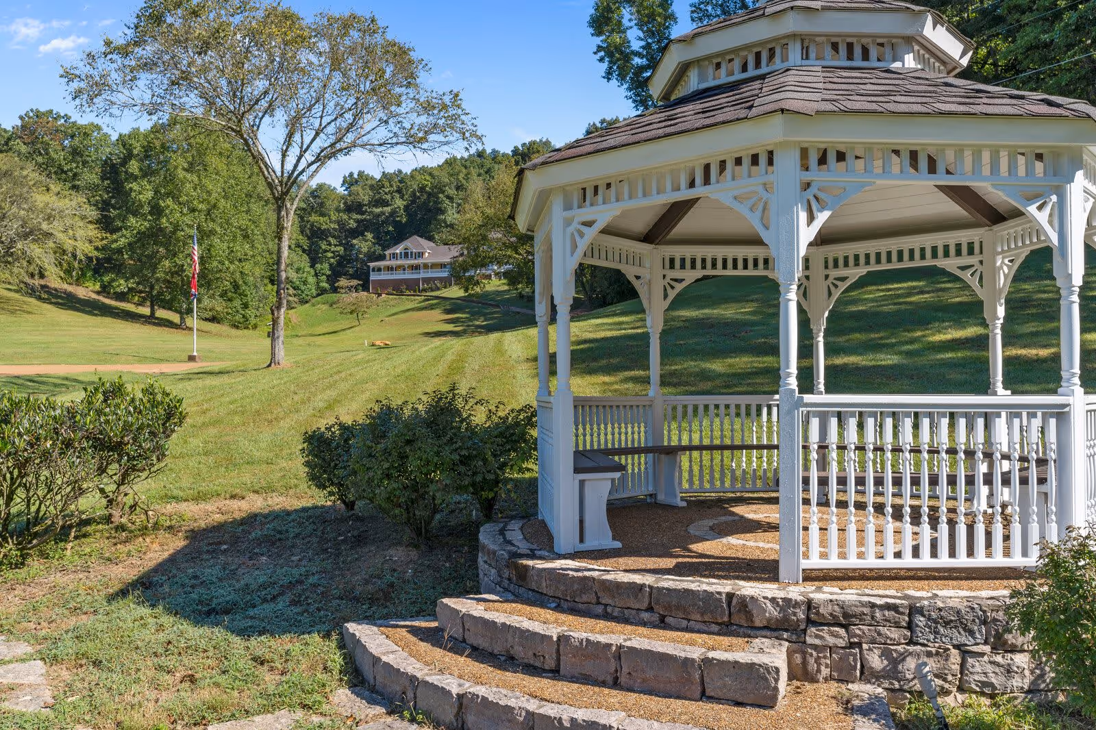 White gazebo on a stone platform overlooking a grassy lawn with trees and a house in the background.