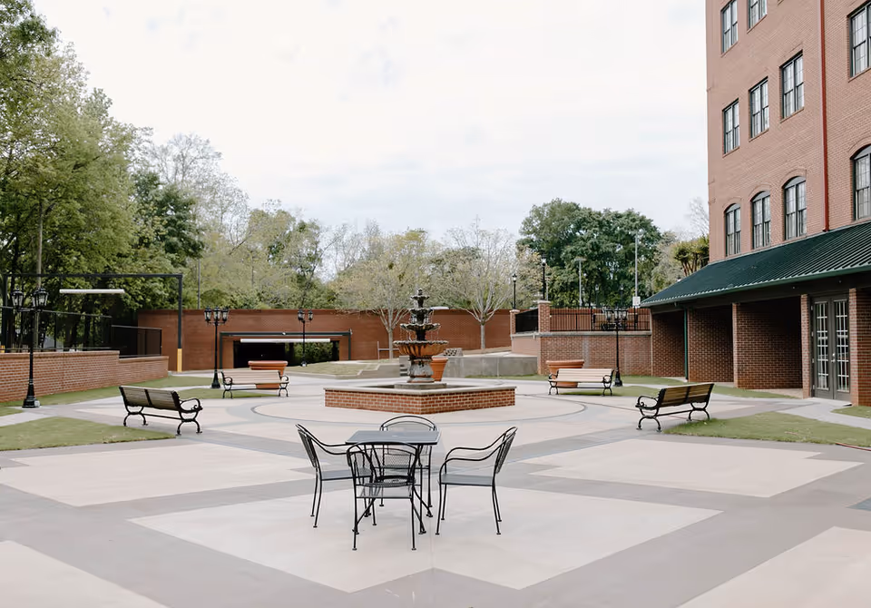 Brick courtyard with a central fountain, outdoor tables and benches in front of a multi-story brick building.