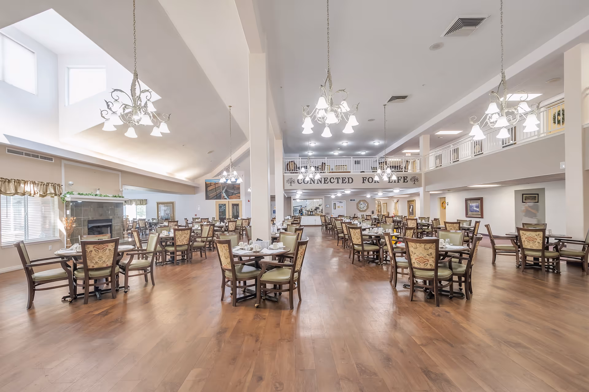 Spacious senior living dining room with multiple set tables, chandeliers, and a mezzanine balcony displaying the sign 'CONNECTED FOR LIFE'.