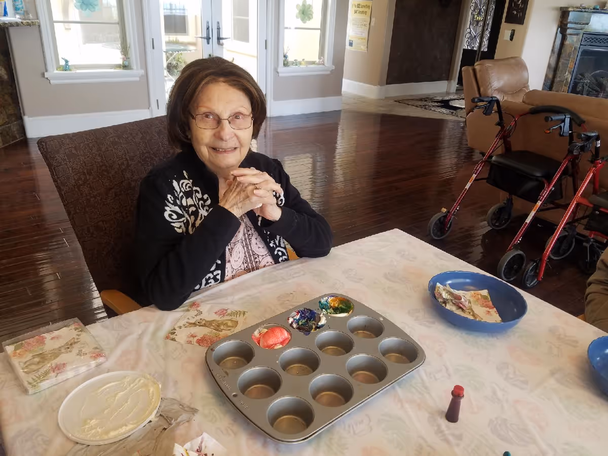 An elderly woman with glasses sitting at a table covered with a floral tablecloth, smiling with her hands clasped. On the table are a muffin tin with colorful paint, a plate with white cream or paint, napkins, a small bottle of red paint, and a blue bowl with a napkin. In the background, there is a living room area with wooden floors, a brown armchair, and two red walkers.