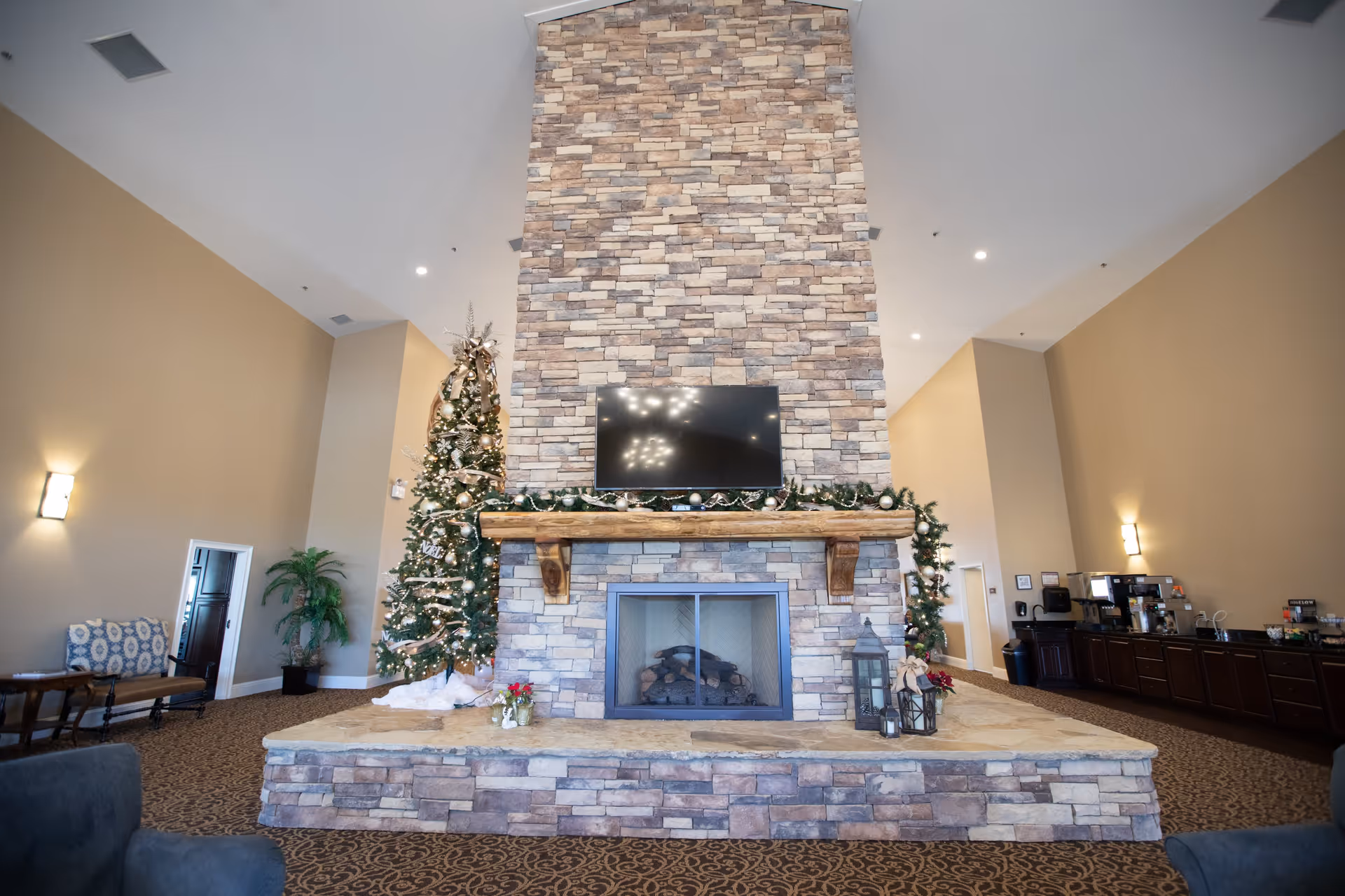 A spacious living area with a tall stone fireplace decorated with garlands and Christmas ornaments. A flat-screen TV is mounted above the fireplace. To the left of the fireplace is a decorated Christmas tree, and to the right is a counter with coffee machines and supplies. The room has beige walls, patterned carpet, and comfortable seating.