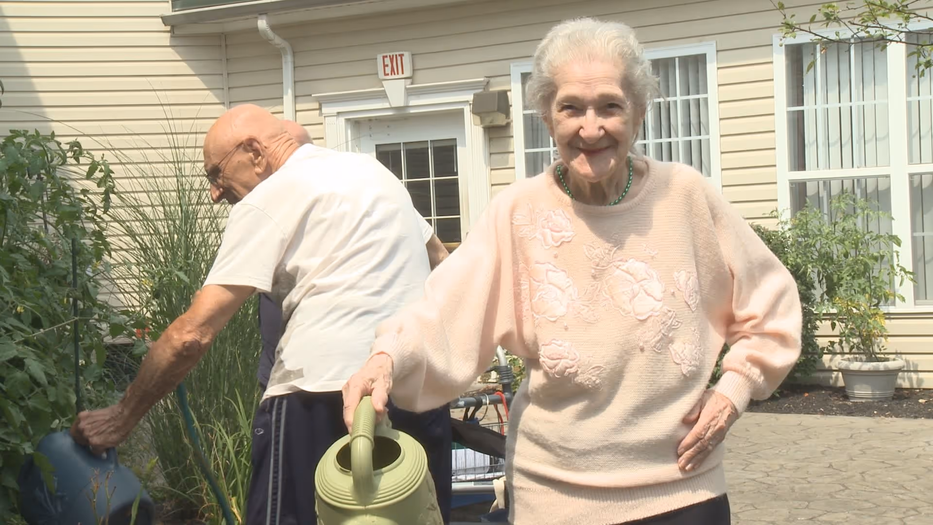 An elderly woman smiling and holding a green watering can in a garden area outside a beige building, with an elderly man watering plants in the background.