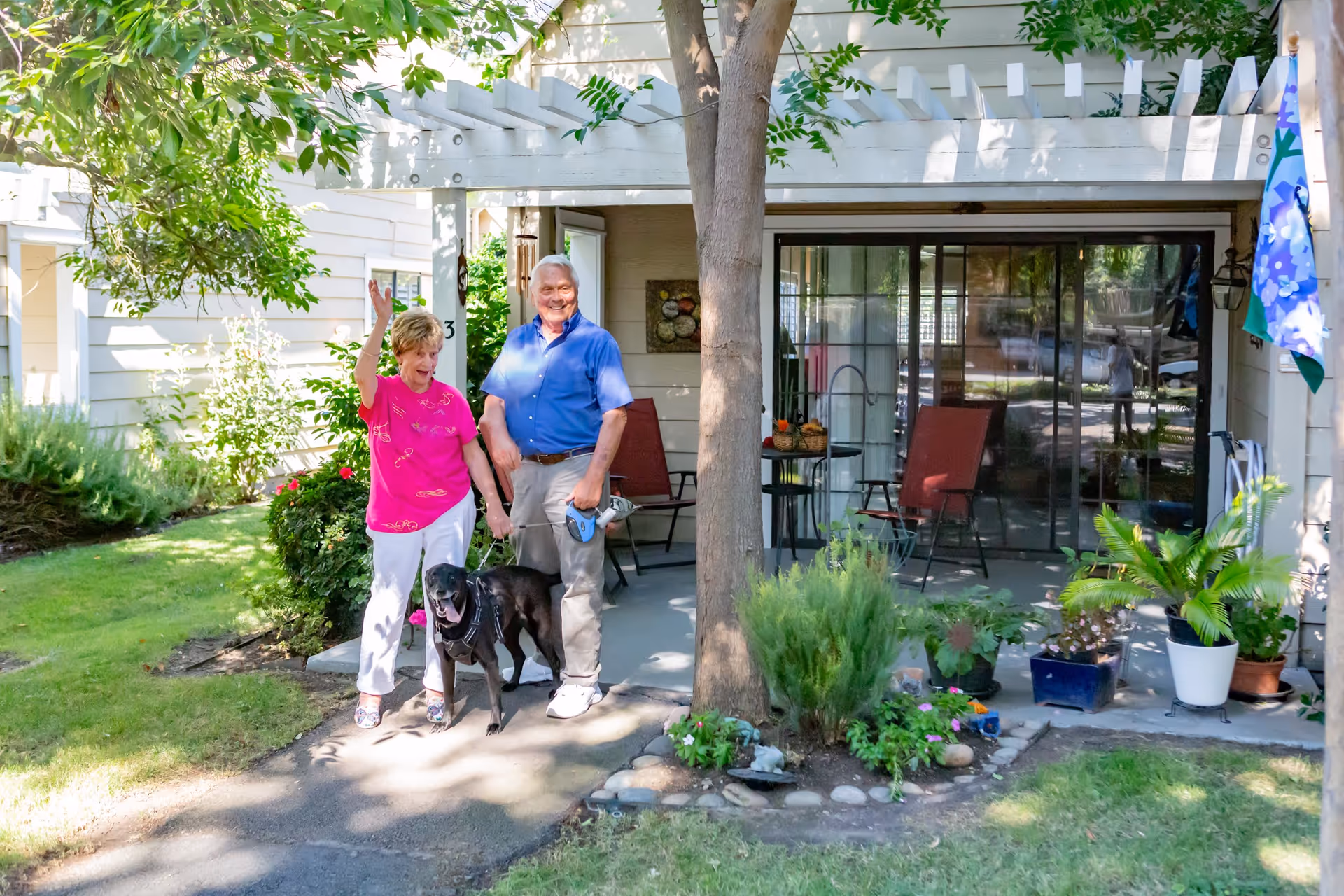 A smiling senior couple standing with a black dog on a path outside a shaded patio of their residence.