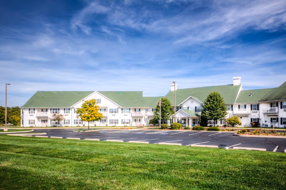 Exterior view of Smith Farms Manor, a two-story senior living facility with white walls and green roofs, surrounded by well-maintained green lawns and trees under a partly cloudy blue sky. The parking lot in front is empty.