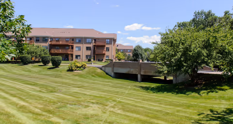 A large grassy lawn with neatly mowed stripes in front of a multi-story brick building with balconies. There are trees and bushes surrounding the lawn, and a concrete bridge or overpass structure is visible on the right side. The sky is clear with a few clouds.