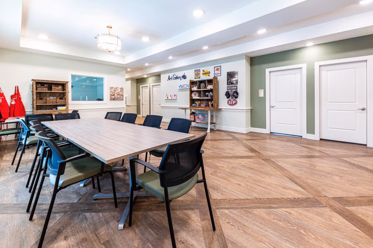 Bright activity room with a long rectangular table surrounded by chairs, shelving labeled 'Art Gallery', and doors on a green-accent wall.