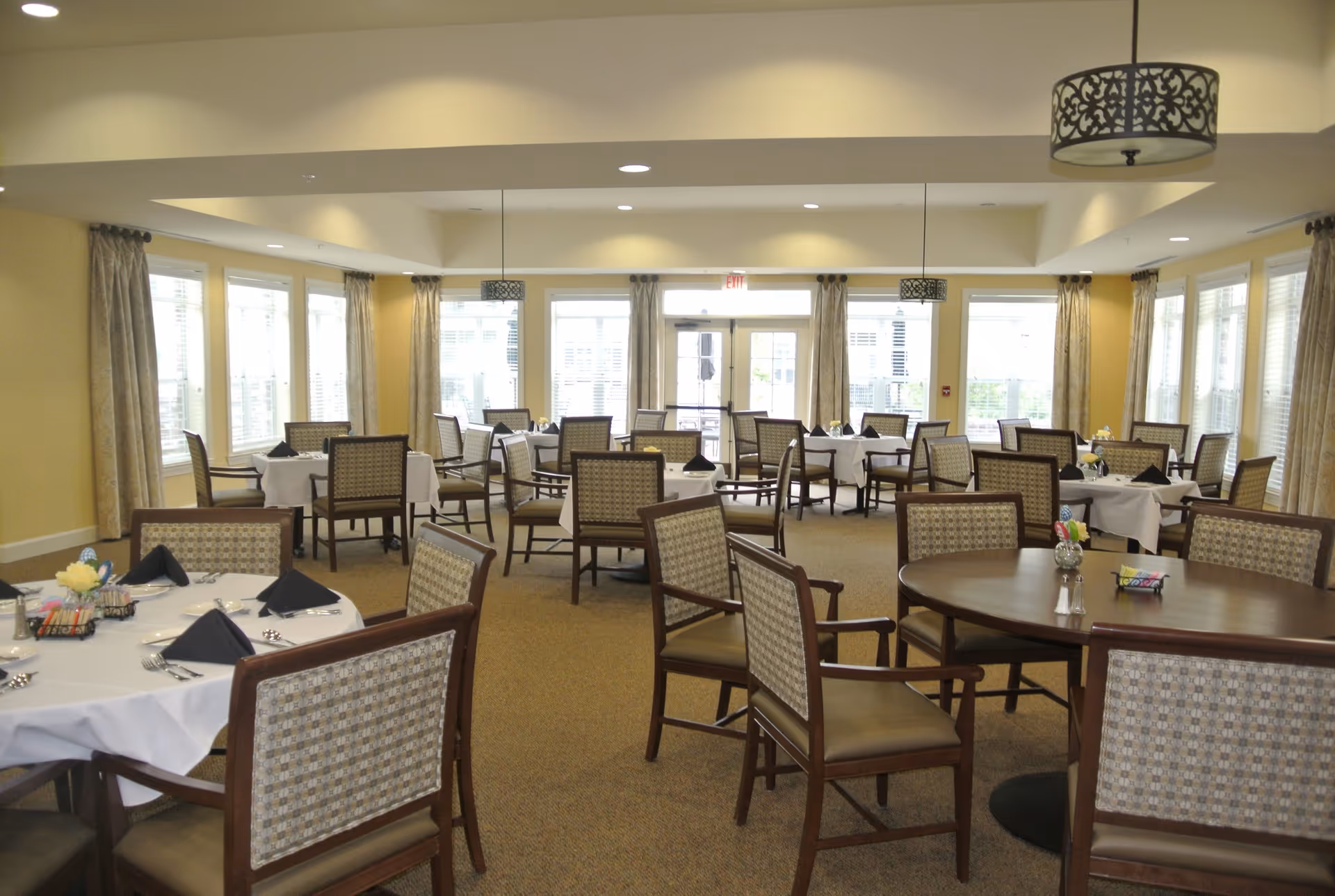 A spacious dining room with multiple round and square tables covered with white tablecloths and set with black napkins, silverware, and small flower arrangements. The room has large windows with beige curtains allowing natural light to fill the space. The ceiling features recessed lighting and decorative hanging light fixtures.