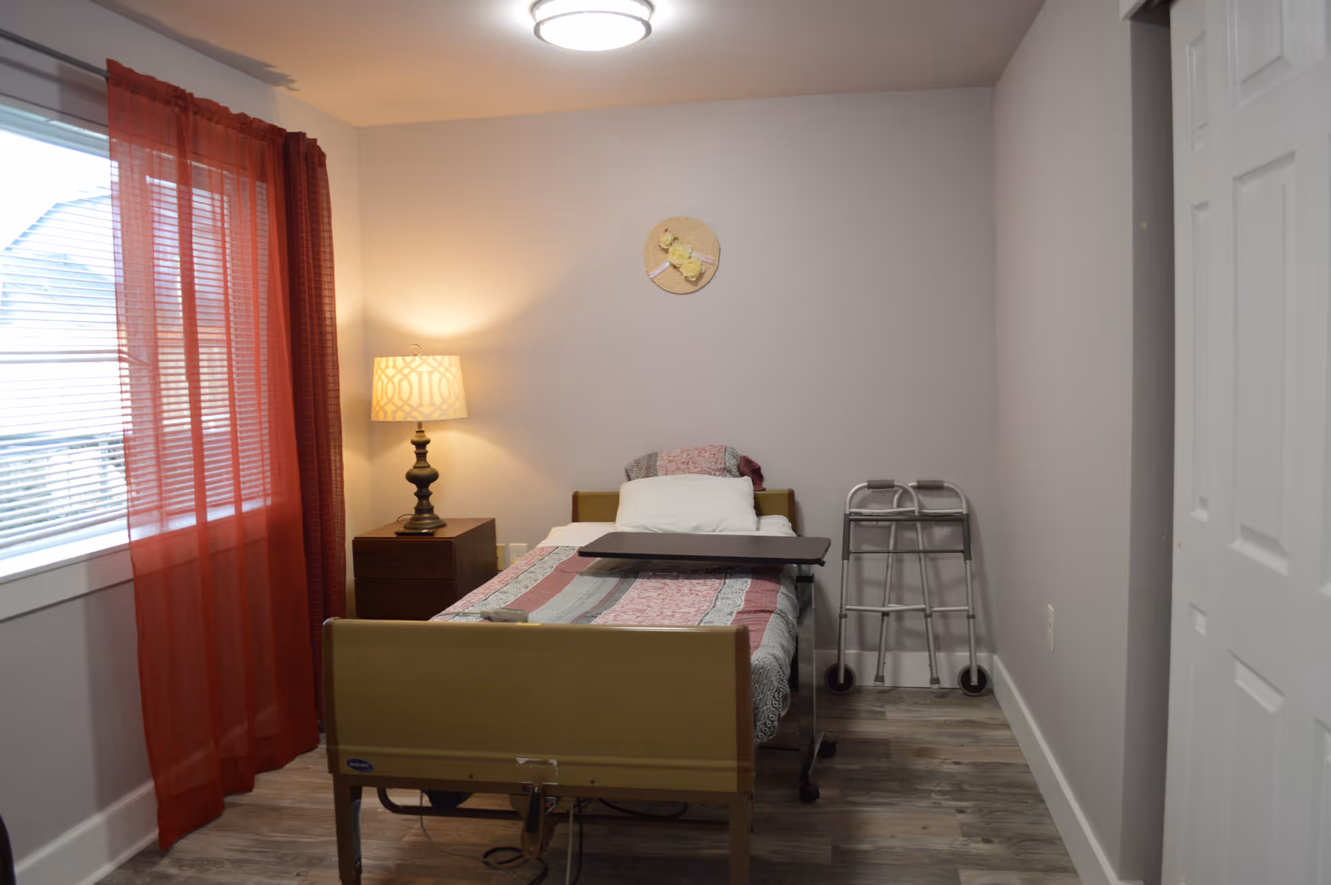A small bedroom with a single bed covered with a patterned quilt and a white pillow. Next to the bed is a wooden nightstand with a decorative lamp. A walker is positioned against the wall near the foot of the bed. The room has light-colored walls, wood flooring, a window with red curtains, and a round wall decoration above the bed.