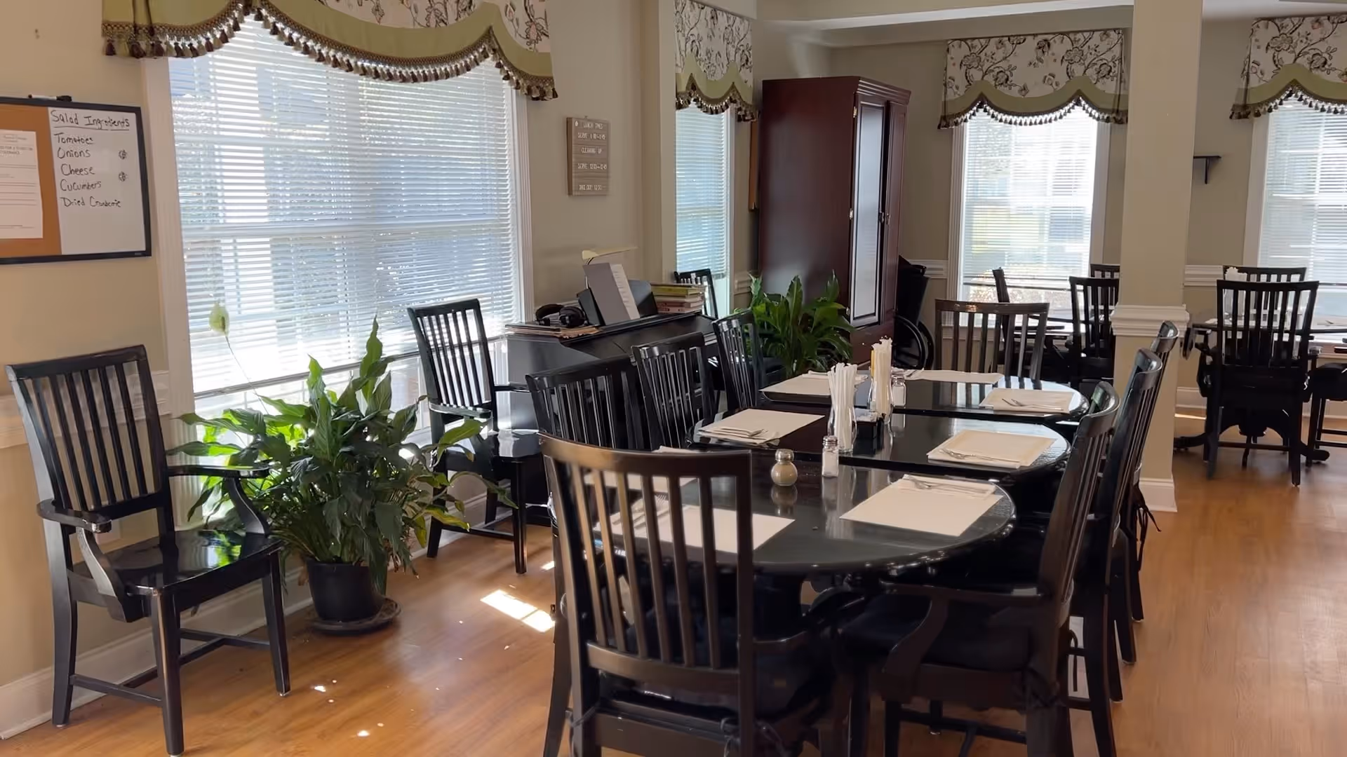 A bright dining room with multiple black wooden tables and chairs arranged neatly. Each table is set with white napkins, utensils, and condiments. Large windows with blinds and decorative valances allow natural light to fill the room. There are green potted plants near the windows and a piano against the wall. The floor is wooden, and the overall atmosphere is clean and inviting.