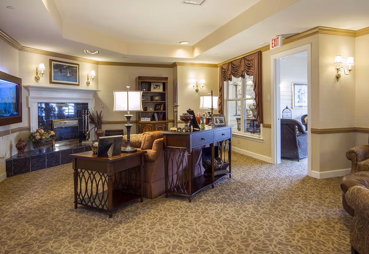 A cozy senior living facility common area with patterned carpet, beige walls, and warm lighting. The room features a brown sofa, wooden side tables with lamps, a fireplace with a painting above it, and a built-in bookshelf. There is a window with draped curtains and an open doorway leading to another room with wicker furniture.