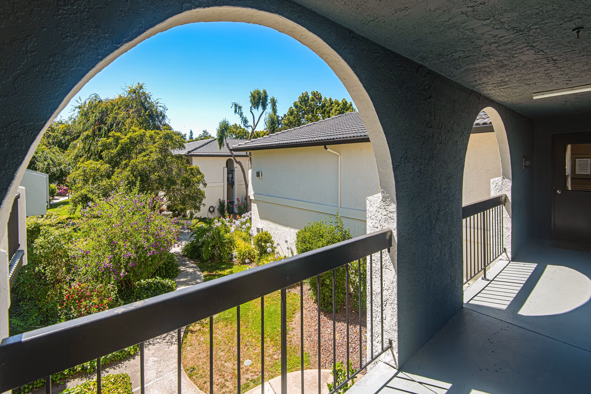 View from a covered outdoor corridor with arched openings overlooking a landscaped garden area with trees, bushes, and a pathway, adjacent to light-colored buildings with tiled roofs under a clear blue sky.