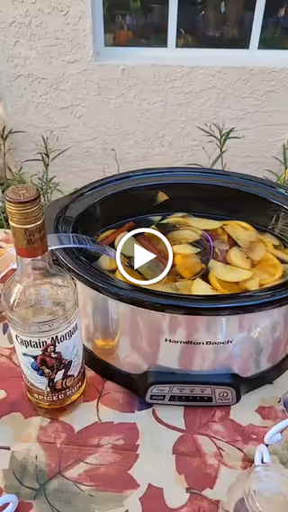 A slow cooker filled with sliced apples and liquid, placed on a table with a floral tablecloth. Next to the slow cooker is a bottle of Captain Morgan Spiced Rum. The setting appears to be outdoors with a beige stucco wall and window in the background.