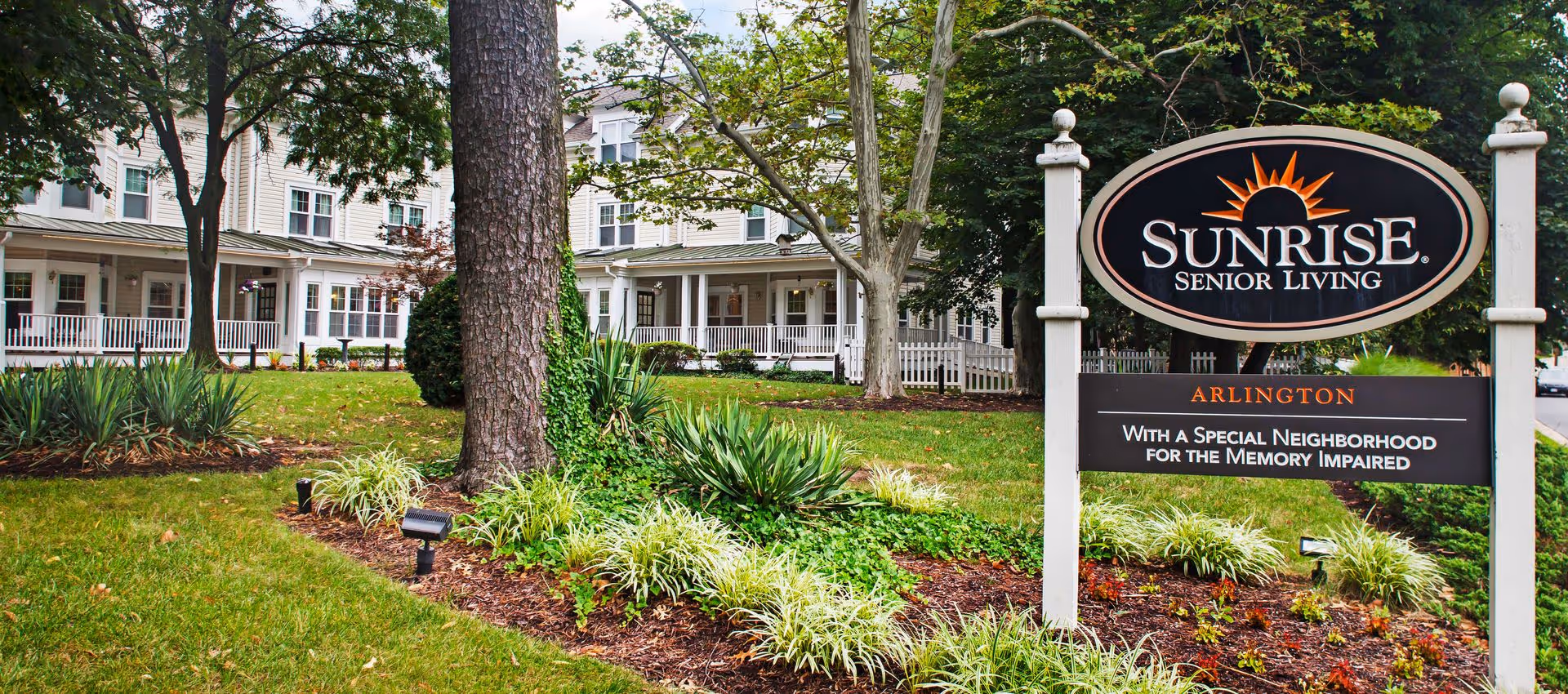 Exterior view of Sunrise Senior Living facility in Arlington with a large sign in the foreground and a multi-story building with white siding and a porch surrounded by trees and landscaped greenery.