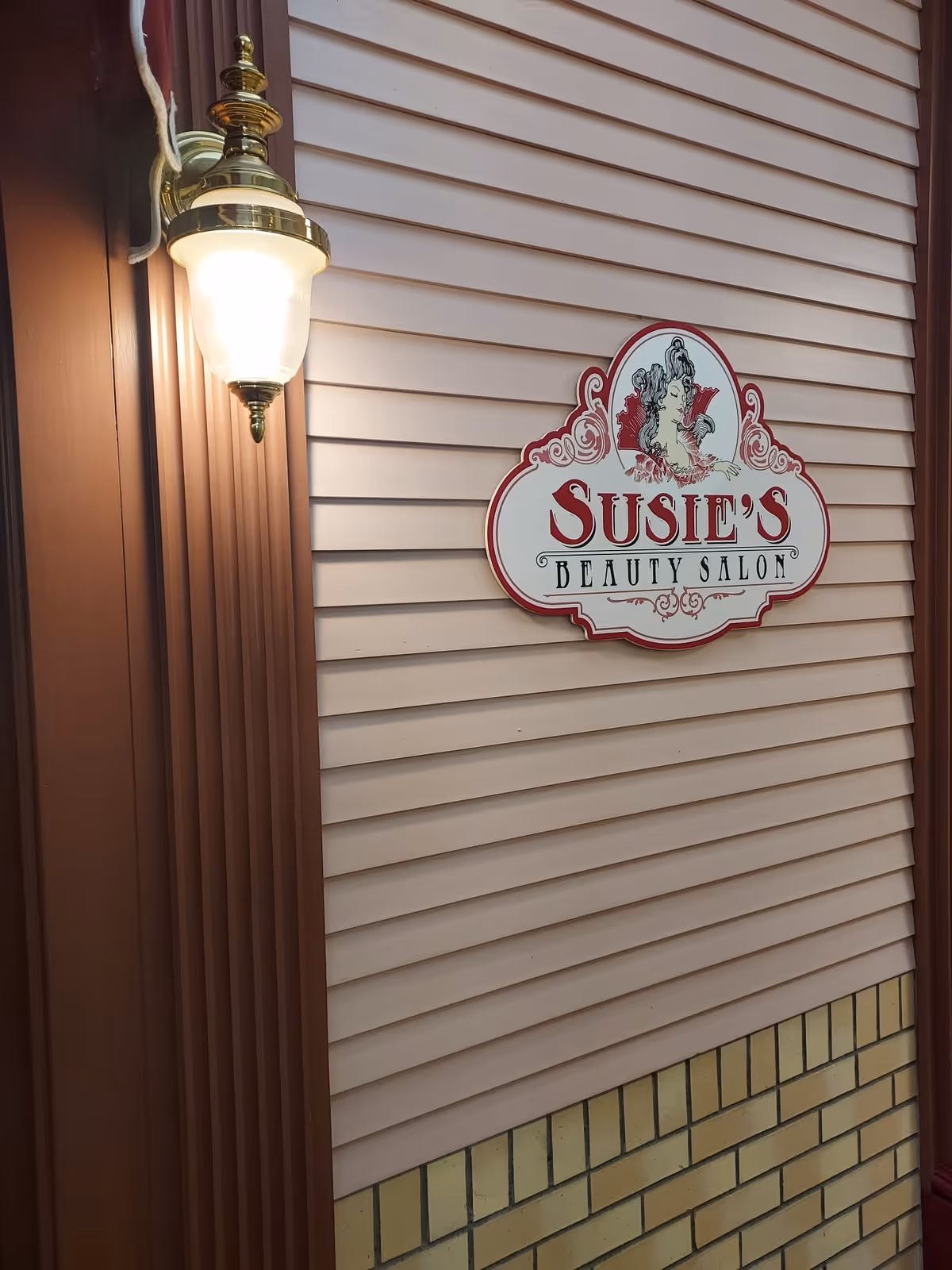 A wall with beige horizontal siding and a lower section of yellow brick. Mounted on the wall is a decorative sign that reads 'Susie's Beauty Salon' with an illustration of a woman with styled hair. To the left of the sign is a brass wall-mounted light fixture with a frosted glass shade that is lit.