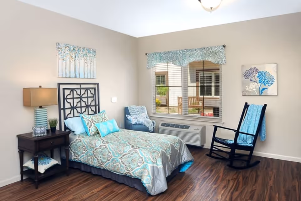 A neatly arranged bedroom with a single bed featuring a patterned blue and gray bedspread and multiple pillows. Next to the bed is a wooden nightstand with a lamp, a small plant, and a framed photo. There is a blue armchair with a white throw blanket near a window with blinds and a blue valance. A black rocking chair with a blue throw is positioned against the wall, which is decorated with two floral paintings. The room has wood flooring and neutral-colored walls.