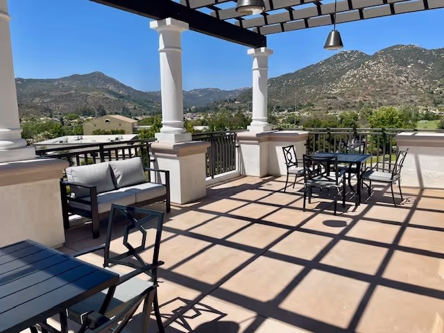 Outdoor patio area with black metal tables and chairs, a cushioned bench, white columns, and a pergola overhead casting shadows on the floor. In the background, there are green trees and hills under a clear blue sky.