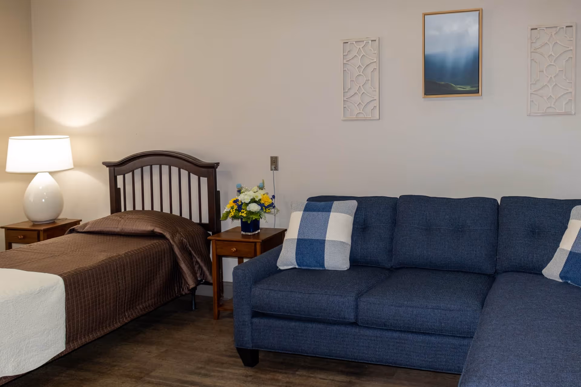 A cozy room featuring a single bed with a brown quilt and pillow, a wooden nightstand with a white lamp and a vase of colorful flowers, and a blue upholstered sofa with checkered pillows. The walls are decorated with two white ornamental panels and a framed picture of a landscape with sunlight breaking through clouds.