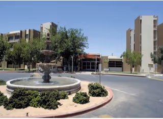 Outdoor view of Kivel Campus of Care featuring a circular fountain with water flowing, surrounded by greenery and bushes, with multi-story buildings in the background under a clear blue sky.