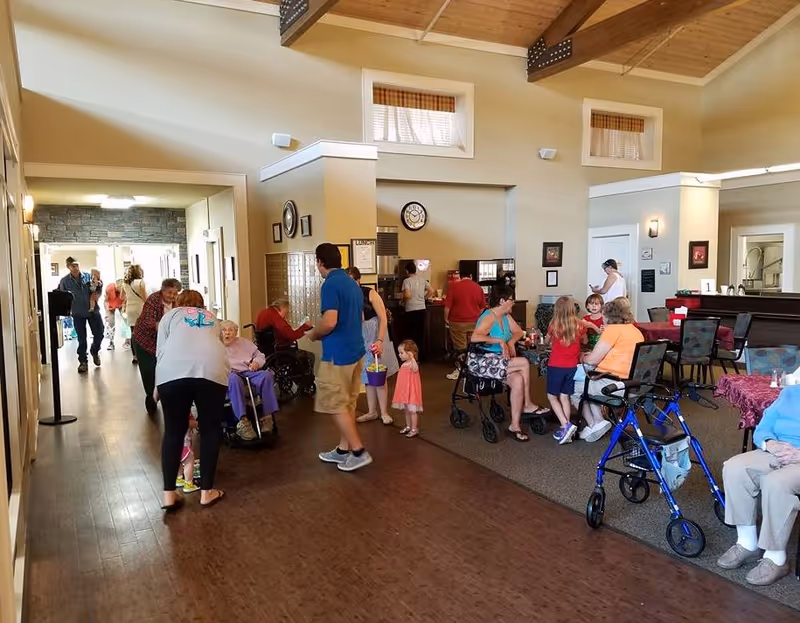 A lively indoor common area at RiverLodge Assisted Living with several elderly residents and visitors interacting. Some people are seated at tables, others are walking or standing, and a few are using walkers or wheelchairs. The room has high ceilings with wooden beams, large windows, and warm lighting.
