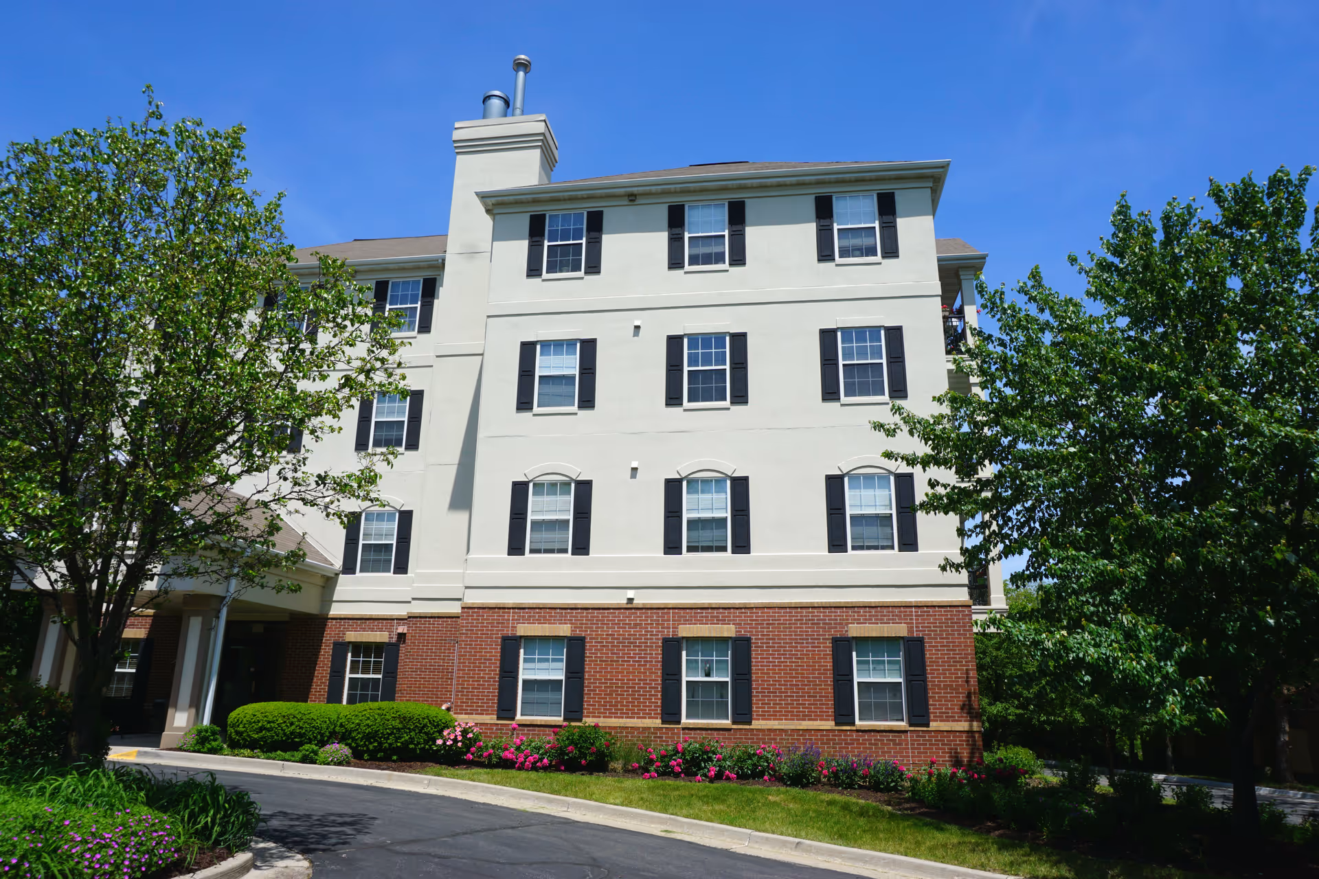 Exterior view of a multi-story retirement facility building with a combination of brick and light-colored siding, surrounded by green trees and landscaping under a clear blue sky.