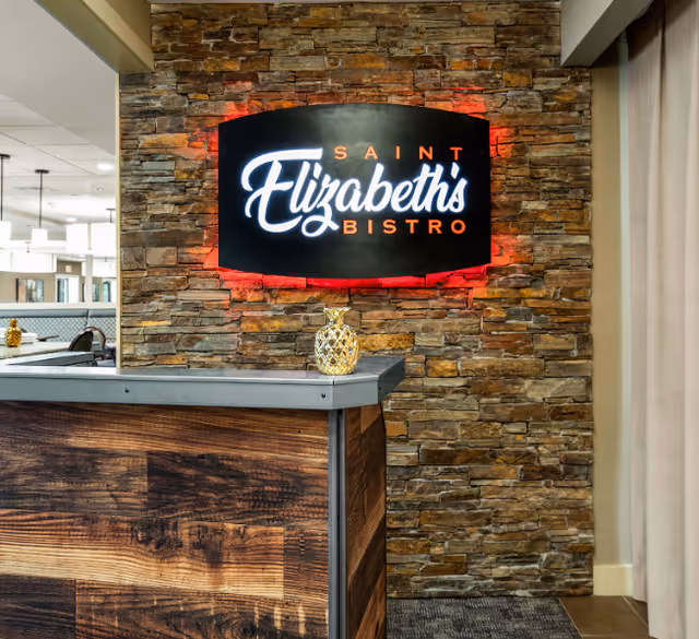 Interior view of a bistro area with a stone wall featuring a lit sign that reads 'Saint Elizabeth's Bistro'. A wooden counter with a decorative pineapple sits in front of the wall, and part of a dining area with tables and chairs is visible in the background.