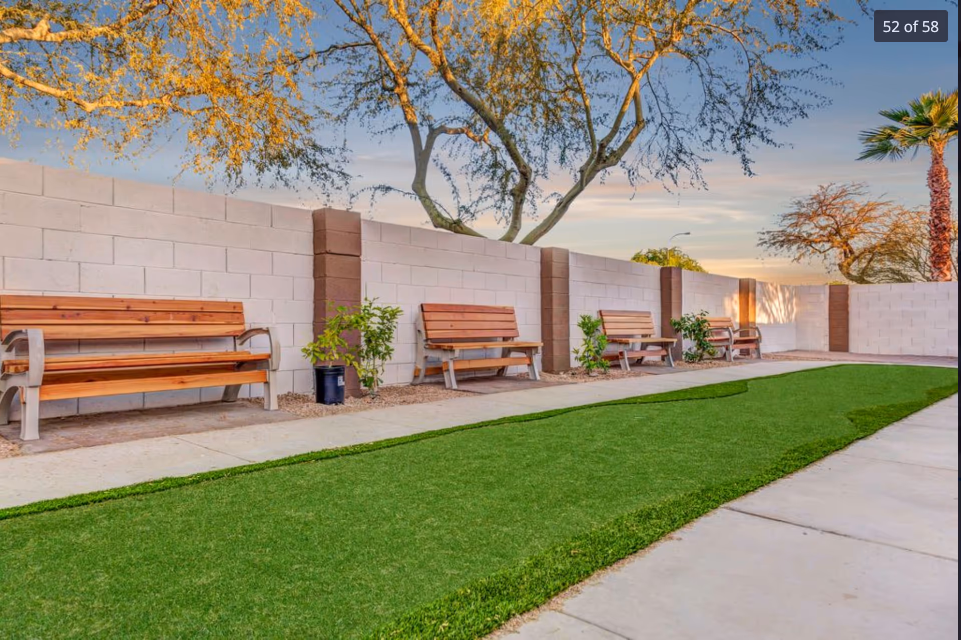 Outdoor area with a row of wooden benches along a white brick wall with brown pillars. There are small plants between the benches and a green artificial turf lawn in front. Trees with sparse leaves and a clear sky are visible in the background.