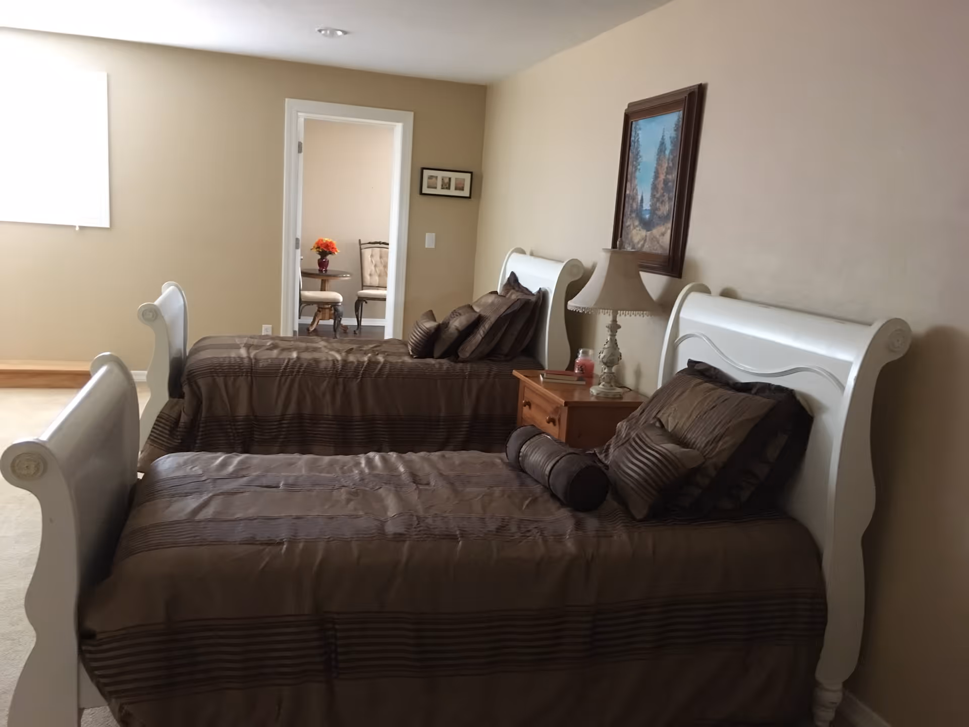 A bedroom with two white wooden twin beds side by side, each made with brown striped bedding and multiple pillows. Between the beds is a wooden nightstand with a decorative lamp and a small candle. A framed landscape painting hangs on the beige wall above the nightstand. In the background, an open doorway reveals a small table with a vase of flowers and two chairs in an adjacent room.