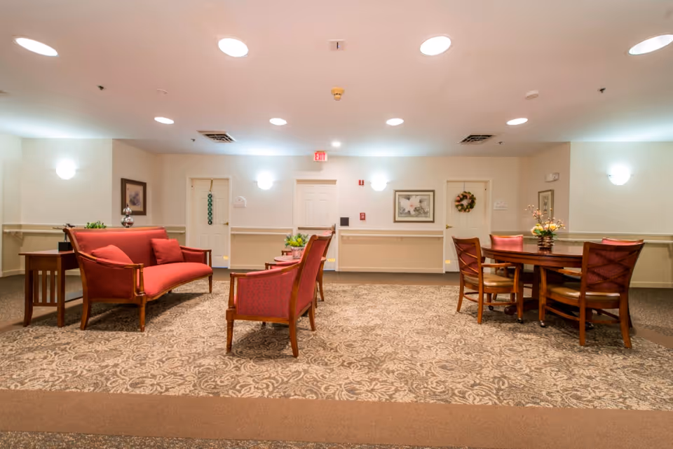 A well-lit common area with red upholstered wooden chairs and a sofa arranged around a patterned carpet. There is a round wooden table with four chairs on the right side, decorated with a flower arrangement. The walls are light-colored with framed artwork and two doors, one adorned with a wreath. Ceiling lights and wall sconces provide illumination.