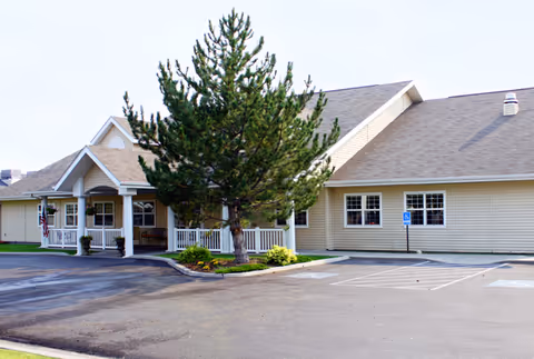 Exterior view of Pomerelle Place Senior Living facility showing a single-story building with beige siding, a covered entrance with white columns, a large tree in front, and a parking lot with marked handicap spaces.