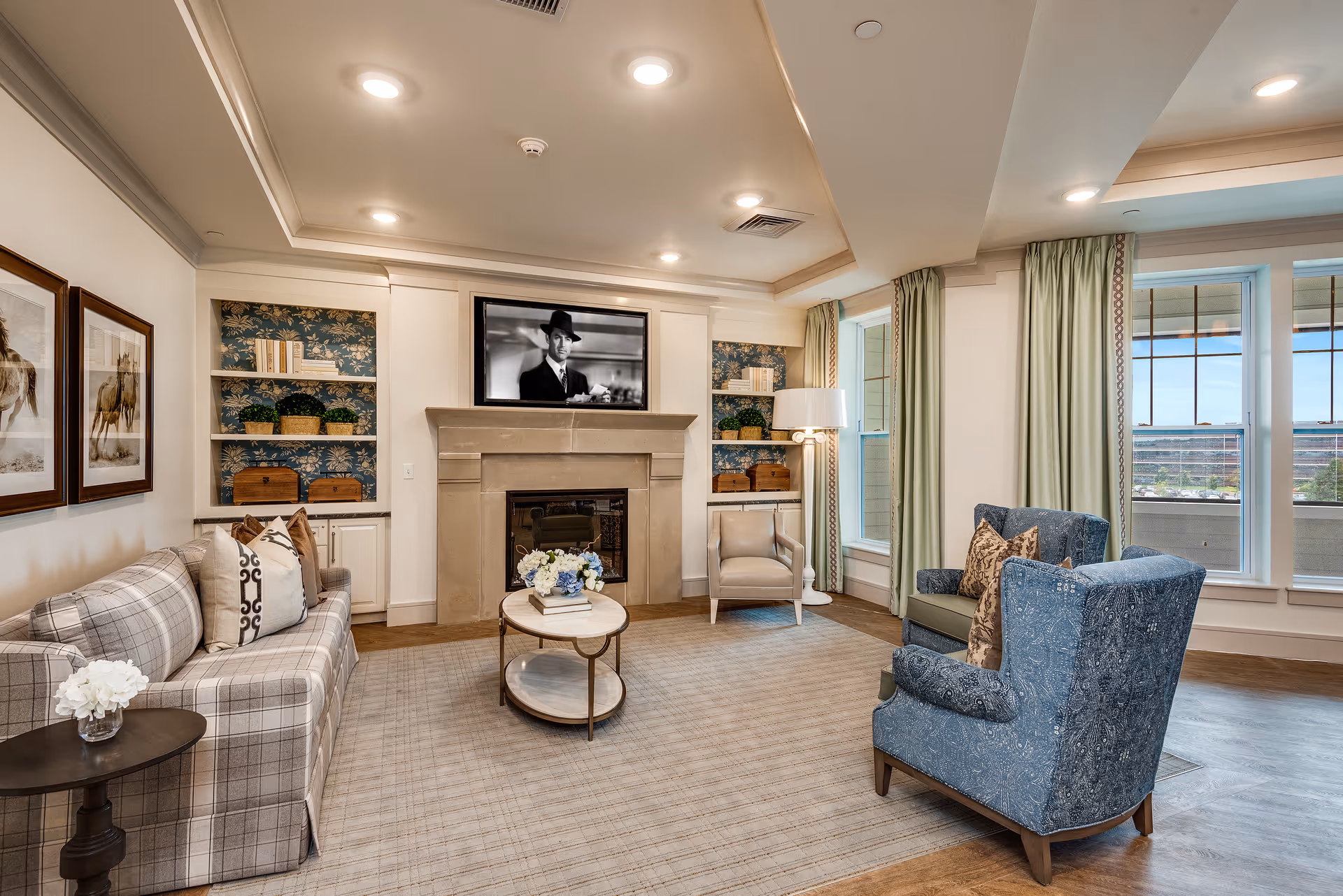 A cozy living room with a plaid sofa, two blue armchairs, a beige armchair, and a round coffee table with flowers. The room features a fireplace with a mounted TV above it, built-in shelves with decorative items, large windows with green curtains, and framed horse artwork on the wall.
