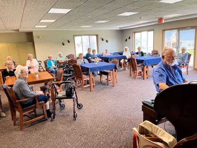 A group of senior residents seated at tables with blue tablecloths in a bright communal dining/activity room, with a man at a piano in the foreground.