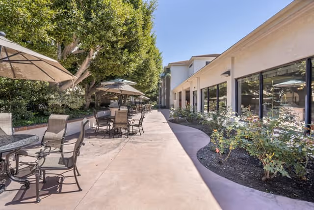 Outdoor patio at a senior living facility with umbrella-shaded tables and chairs along a walkway next to the building and landscaping.