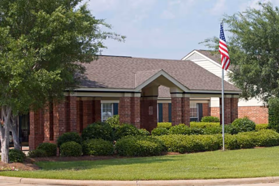 Exterior view of a single-story brick building with a gabled roof, surrounded by green bushes and trees. An American flag is displayed on a flagpole in front of the building under a partly cloudy sky.