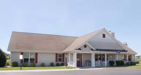 Single-story building with beige siding and a brown roof, featuring a covered front porch with white columns and several chairs. The building is surrounded by a paved driveway and small bushes, under a clear blue sky.
