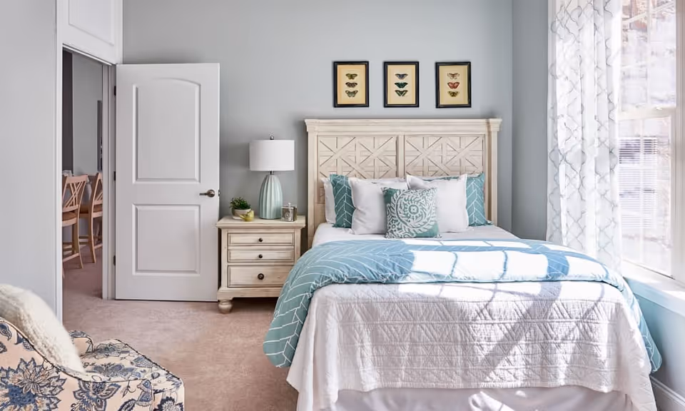 A bright bedroom with a bed featuring a white quilt and teal patterned pillows and blanket. Above the bed are three framed butterfly prints. To the left of the bed is a nightstand with a lamp, a small plant, and a clock. A door is open, showing a glimpse of a dining area with wooden chairs. A large window with patterned curtains lets in natural light on the right side of the room. A patterned armchair is partially visible in the foreground.