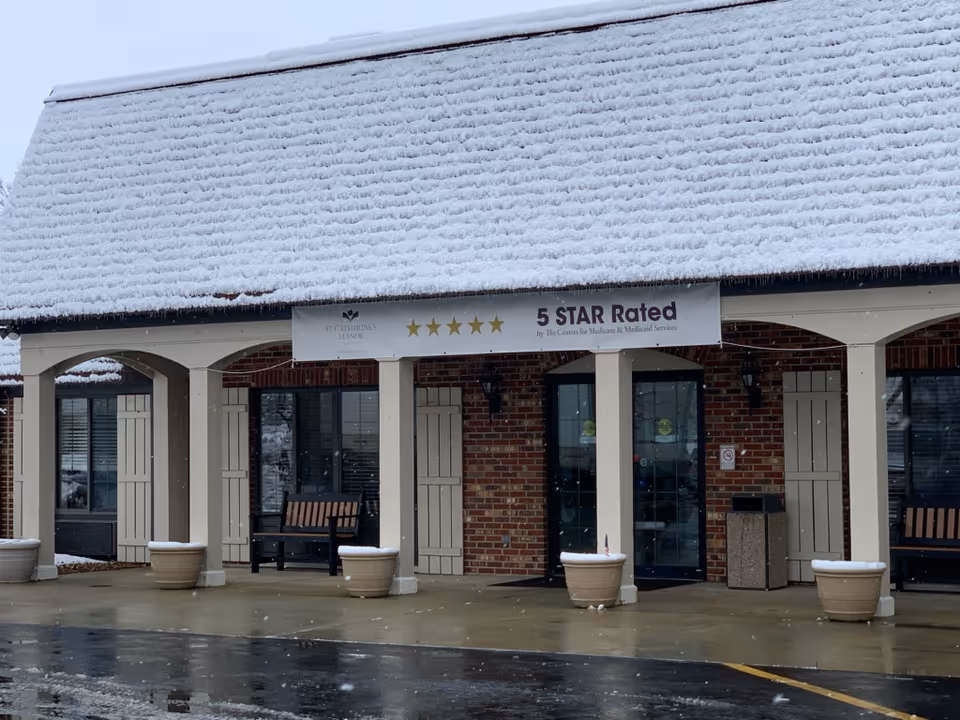 Exterior view of a brick building with a snow-covered roof and a covered entrance supported by white columns. There are benches and large flower pots near the entrance. A banner above the entrance reads '5 STAR Rated by The Centers for Medicare & Medicaid Services.'