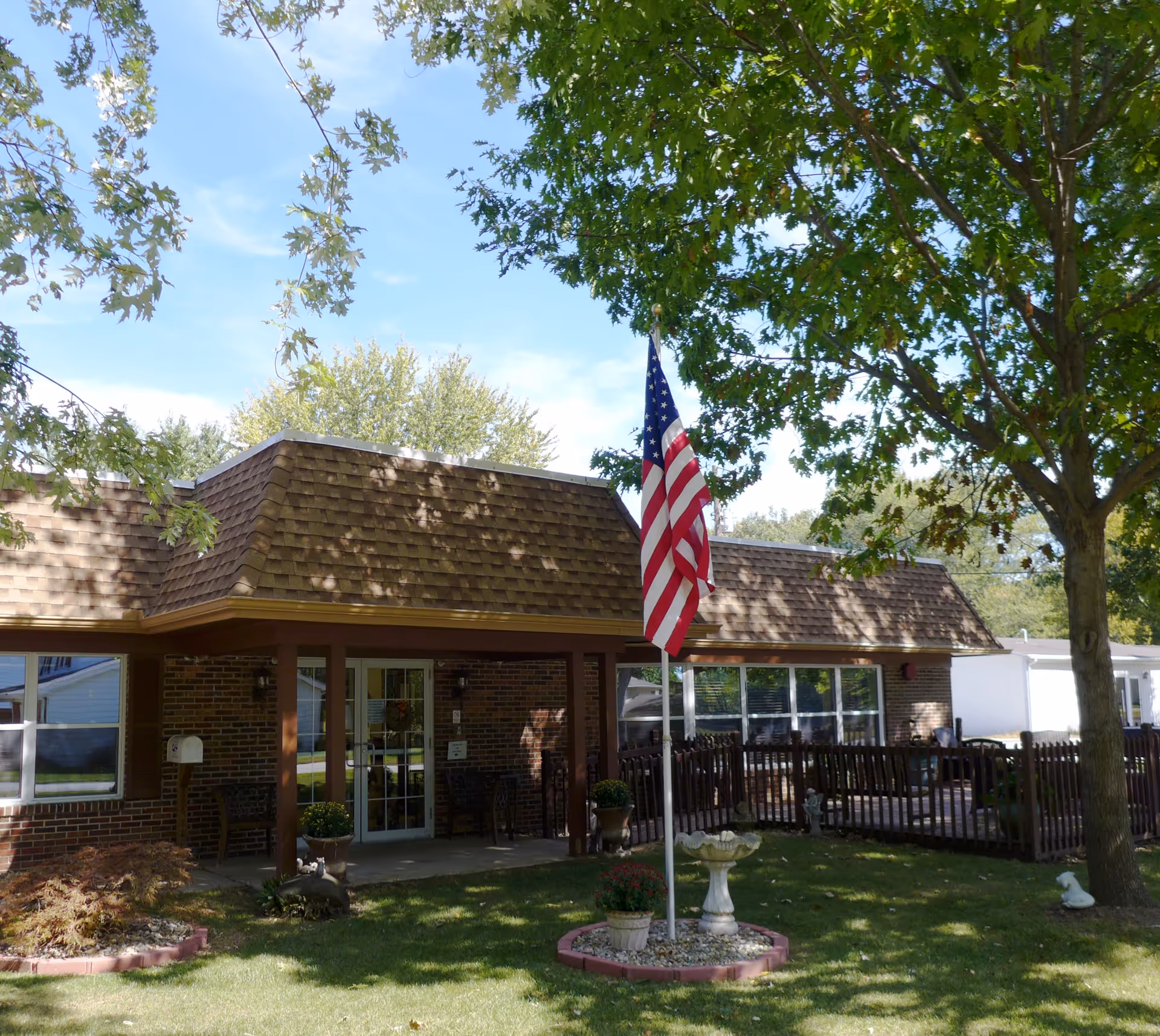 Exterior view of a single-story brick building with a brown shingled roof, an American flag on a flagpole in front, and a small garden area with a birdbath and potted plants. There are trees providing shade and a fenced patio area to the right.