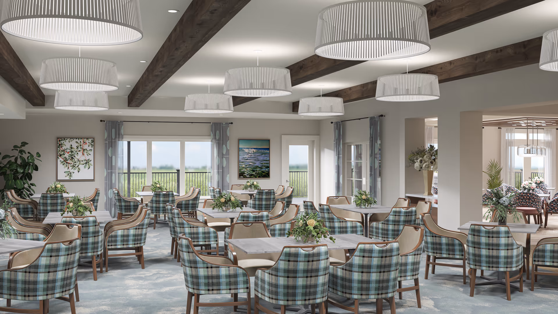 A spacious and well-lit dining room with multiple tables and chairs arranged neatly. The chairs have a blue and green plaid pattern with wooden legs. The ceiling features wooden beams and several large, modern pendant lights. The room has large windows and glass doors letting in natural light, with curtains on either side. There are framed artworks on the walls and floral centerpieces on each table, creating a welcoming and comfortable atmosphere.