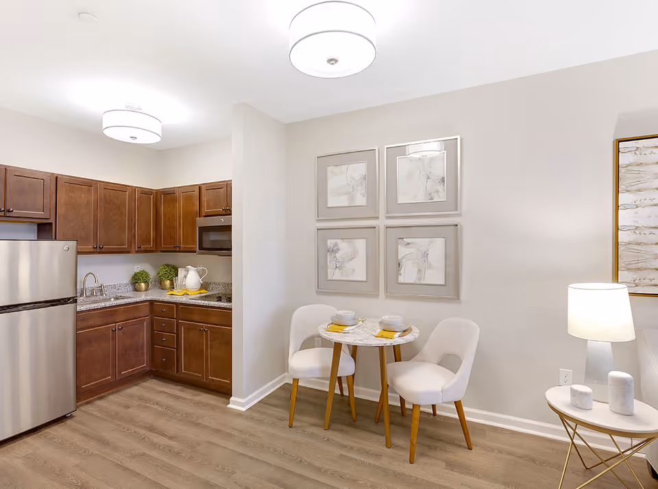 Small open kitchen with stainless steel refrigerator and dark wood cabinets next to a round bistro table and two upholstered chairs against a light-colored wall.