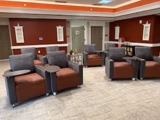A seating area with multiple comfortable armchairs arranged in pairs, each pair sharing a small table. The room has red walls with white trim, a beige carpeted floor, and recessed lighting in the ceiling. There are doors and wall decorations visible in the background.