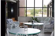 Sunlit common area with round tables and wicker chairs by large windows overlooking outdoor greenery.