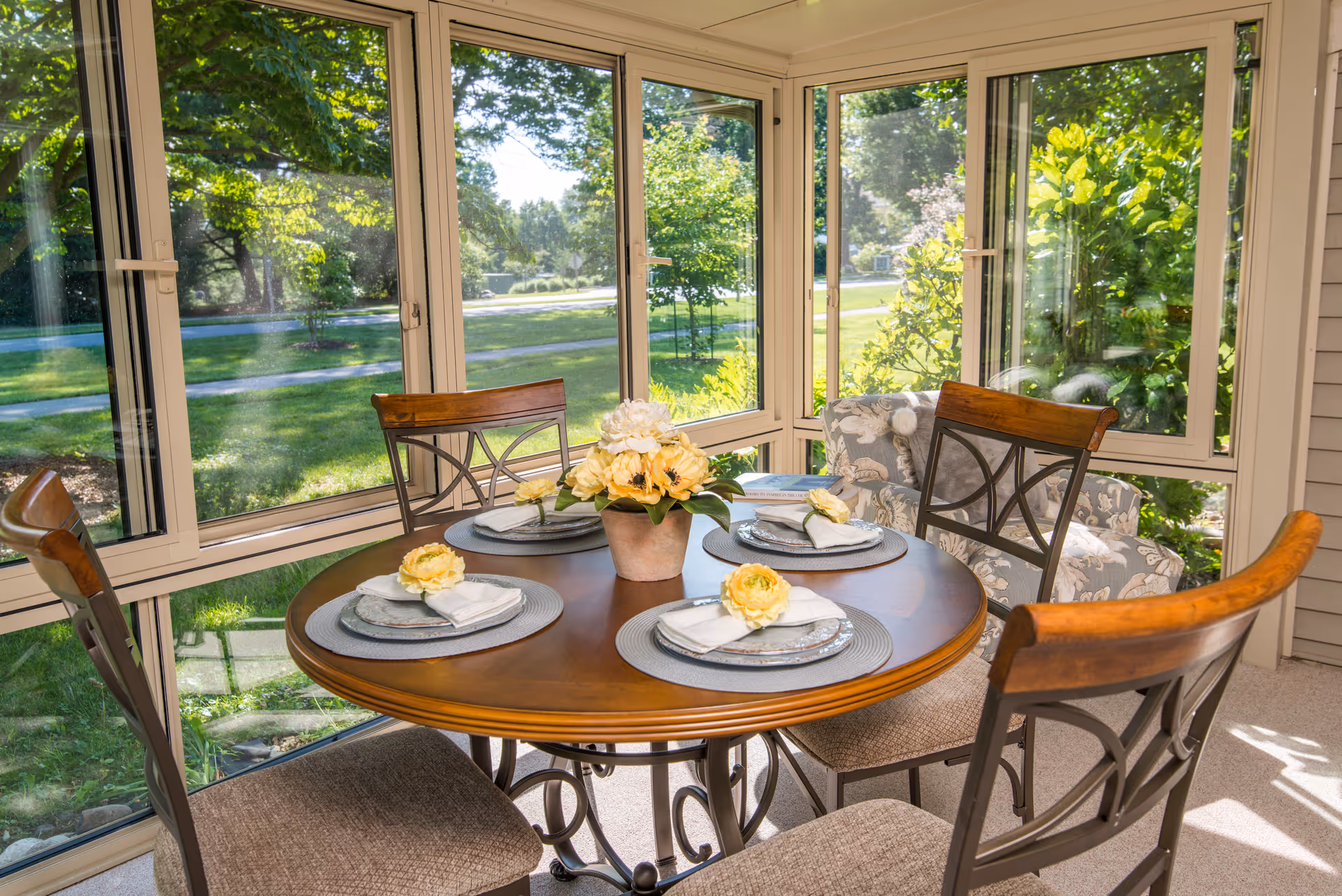 Sunlit dining nook with a round wooden table set for four and large windows overlooking a green yard.