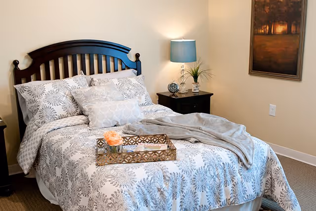 A neatly made bed with a dark wooden headboard in a senior living facility bedroom. The bed has white and gray patterned bedding with multiple pillows and a gray throw blanket. A decorative tray with a flower and magazines is placed on the bed. Next to the bed is a dark wooden nightstand with a blue lampshade, a small clock, and a plant. A framed painting hangs on the beige wall above the nightstand.