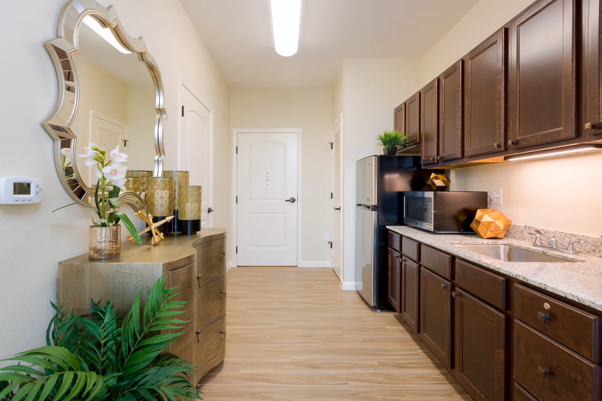 A bright kitchen area with wooden cabinets, a granite countertop, a stainless steel sink, microwave, and refrigerator. On the left side, there is a decorative console table with a large ornate mirror above it, some decorative items, and a potted plant below. The floor is light wood, and the walls are painted a light cream color.